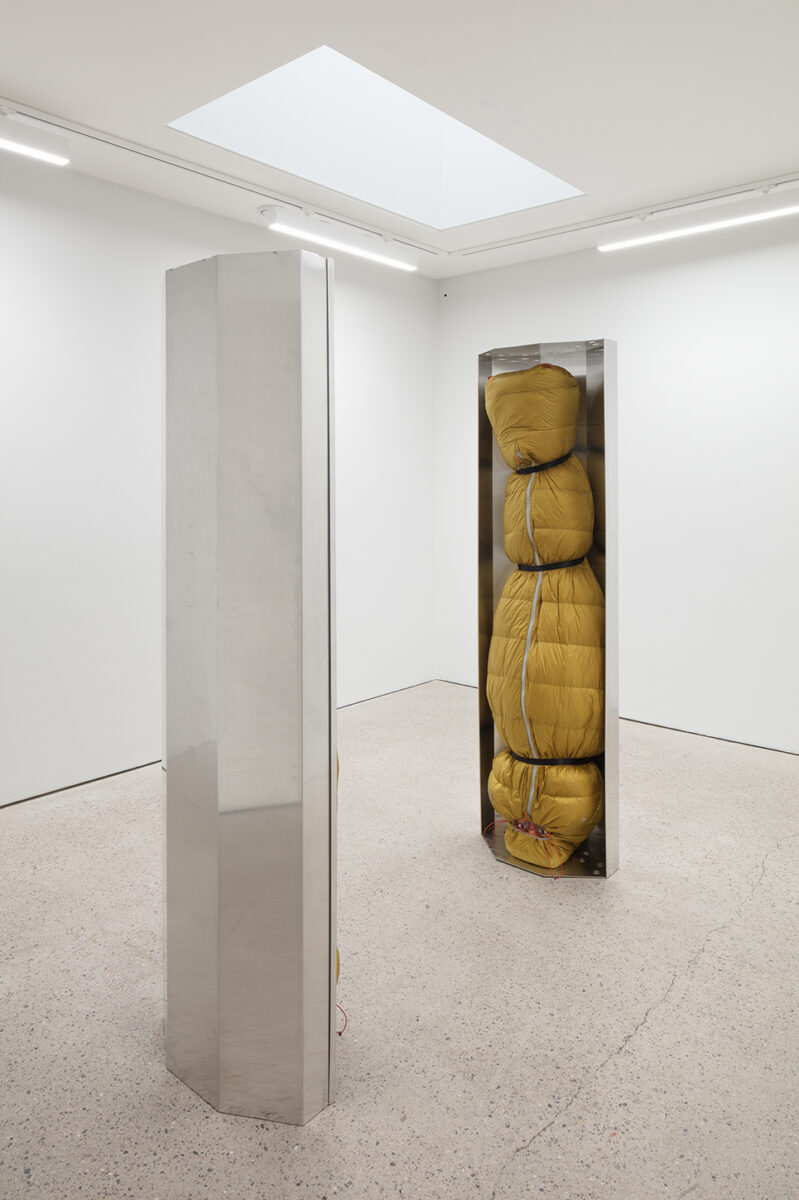 Human-sized figures, wrapped in green thermal sleeping bags and suspended upside down inside stainless-steel trays.