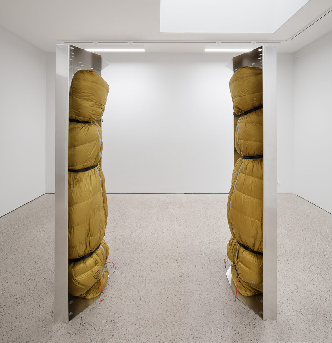 Human-sized figures, wrapped in green thermal sleeping bags and suspended upside down inside stainless-steel trays.